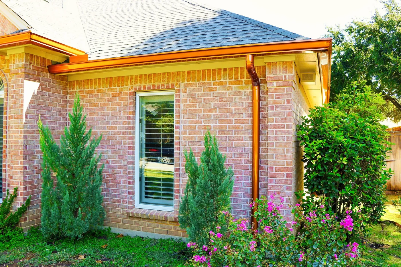 A copper-tone gutter and downspout beside a window on a red brick home