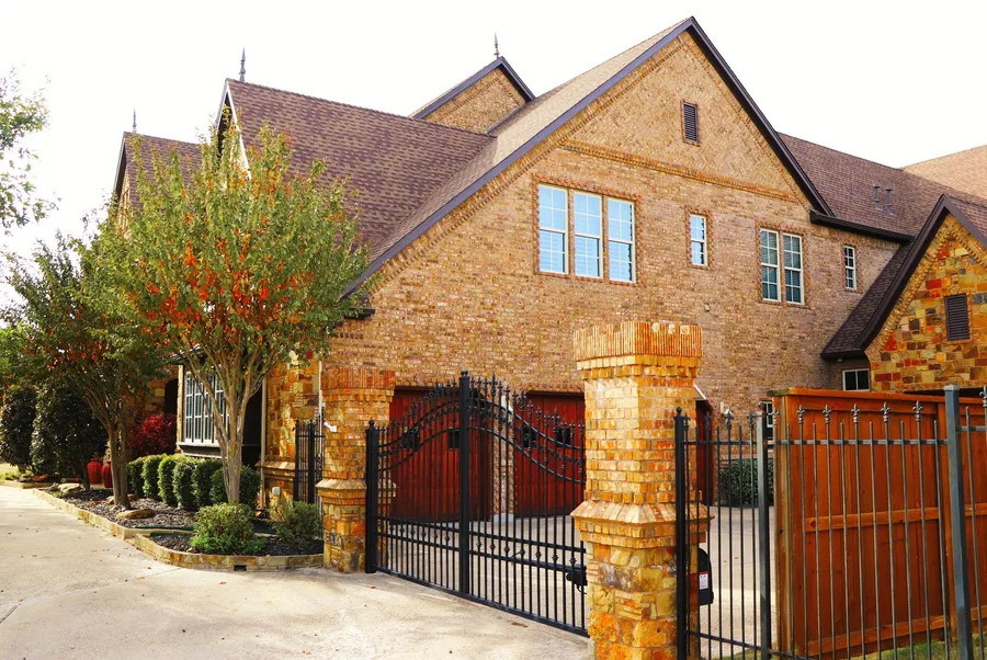 A brick and stone two-story with a brown shingle roof behind an iron driveway gate
