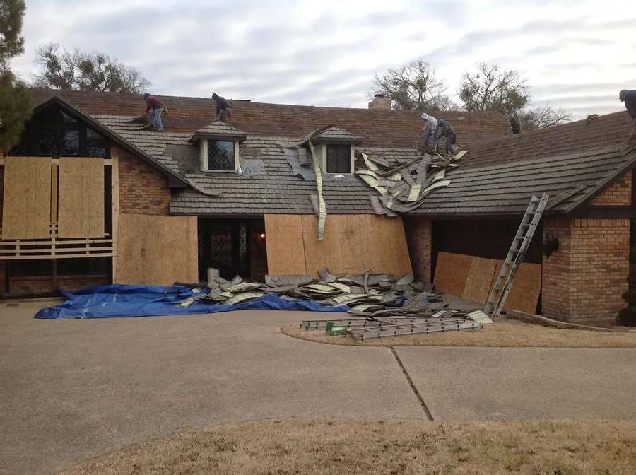 A crew tearing off the roof of a brick home, with the windows boarded